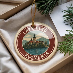 May include: Round ceramic ornament featuring a scenic illustration of Lake Bled, Slovenia. The design includes a church, lake, and mountains, with text reading "Lake Bled Slovenia" in a circular border. The ornament is in a gift box.