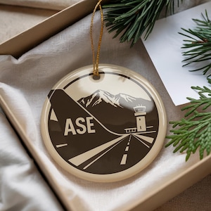 May include: Round Christmas ornament with a tan background and a brown graphic of a mountain, airport runway, and control tower. The letters "ASE" are printed on the left side of the ornament. A gold string is attached for hanging.