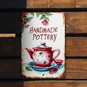May include: A white metal sign with a red and pink teapot illustration and the text "HANDMADE POTTERY" in red lettering.