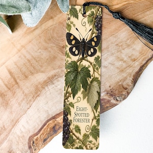 May include: A rectangular bookmark featuring a black butterfly with yellow spots, green grape leaves, and dark purple grapes on a cream background. The text Eight-Spotted Forester is printed in black. A black tassel is attached to the top hole.