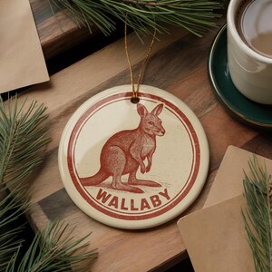 May include: A round, cream-colored ornament with a red illustration of a wallaby. The word "WALLABY" is printed in red at the bottom. The ornament has a gold string for hanging. The background includes a wooden surface and greenery.