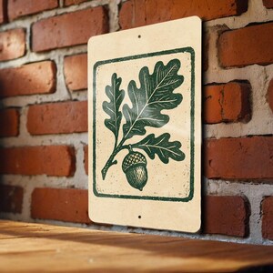 May include: A rectangular metal sign featuring a green oak leaf and acorn design within a green border. The sign has a cream-colored background and is mounted on a brick wall. The sign is a decorative item.