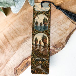 May include: A rectangular bookmark featuring two stacked circular illustrations of the Bay of Fundy in Nova Scotia, surrounded by a decorative green leaf border. The bookmark includes a black tassel at the top and the text Bay of Fundy Nova Scotia.