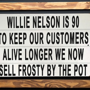 May include: A white sign with black text on a wooden background. The sign reads "Willie Nelson is 90. To keep our customers alive longer we now sell frosty by the pot."