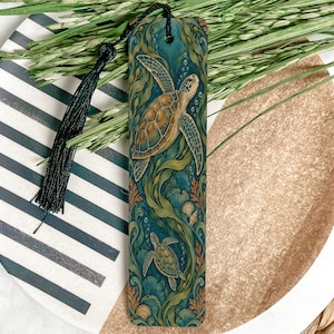 May include: A rectangular bookmark featuring a detailed illustration of two sea turtles swimming among green seaweed and coral in deep blue water. The bookmark has a black tassel attached to the top through a small hole at the center.