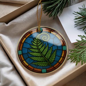 May include: Round ceramic ornament with a stained-glass design featuring a green fern frond against a blue background. The ornament has a gold-colored trim and a gold string for hanging. It is presented in a gift box.