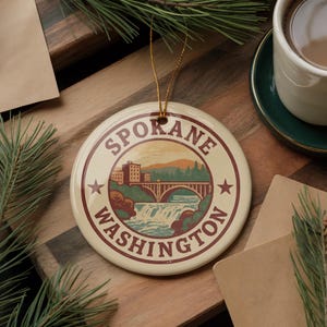 May include: Round ceramic ornament featuring a Spokane, Washington design. The ornament has a cream-colored background with a circular illustration of a bridge, waterfall, and buildings. The words "Spokane Washington" are printed around the illustration in a burgundy color.
