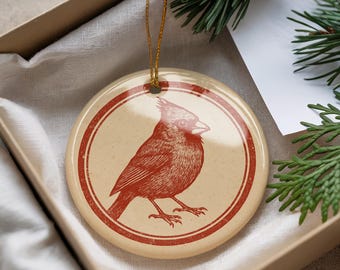 May include: Round ceramic ornament featuring a red cardinal bird illustration within a red double-lined circle. The ornament has a beige background and a gold string for hanging. The ornament is packaged in a box.