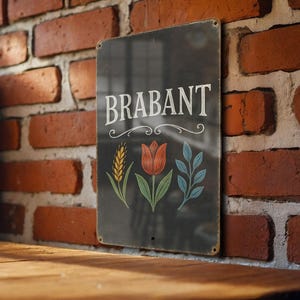 May include: A rectangular black metal sign with the word "BRABANT" in white script. The sign features illustrations of wheat, a tulip, and leaves. The sign is mounted on a red brick wall.