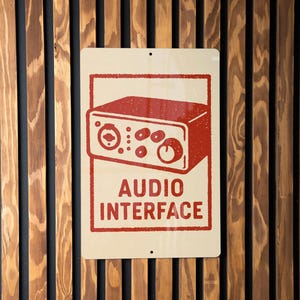 May include: A rectangular sign with a cream background and a red illustration of an audio interface. The sign reads "AUDIO INTERFACE" in red block letters. The sign is mounted on a wooden wall.