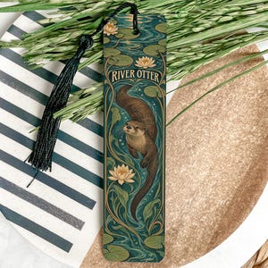 May include: A rectangular bookmark featuring a brown river otter swimming in a teal pond with green lily pads and pale yellow water lilies. The text RIVER OTTER is printed in white at the top. A black tassel is attached to the top with a cord.