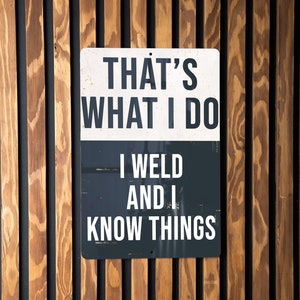 May include: A black and white metal sign with the text "That's what I do. I weld and I know things." The sign is hanging on a wood slat wall.