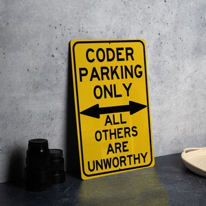 May include: Yellow metal sign with black text that reads "Coder Parking Only" with arrows pointing in both directions. The text below the arrows reads "All Others Are Unworthy".