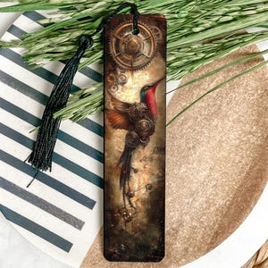 May include: A steampunk-themed bookmark featuring a hummingbird with gears and cogs. The bird has red, brown, and orange feathers. A black tassel hangs from the top. The bookmark is on a striped plate with greenery.