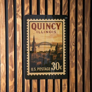 A vintage-style postage stamp from the United States, featuring an illustration of a cityscape and a bridge, hanging on a wooden wall.
