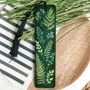 May include: A green bookmark with a black tassel featuring a watercolor illustration of various green leaves and ferns.