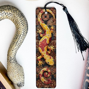 May include: A decorative bookmark featuring a lizard design in shades of yellow, red, and brown, with a black tassel. The bookmark is placed near a decorative snake and a book with a dark cover.