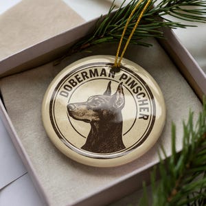 May include: A round, cream-colored ornament featuring a Doberman Pinscher illustration in brown. The dog's profile is centered within a circular frame, with the words "Doberman Pinscher" encircling it. The ornament is in a gift box with a gold string.