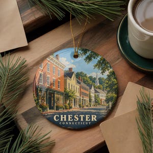 May include: A round ceramic ornament featuring a printed illustration of a street scene in Chester, Connecticut, with historic brick and yellow buildings, green trees, and a church steeple. The text CHESTER CONNECTICUT is printed in white at the bottom.