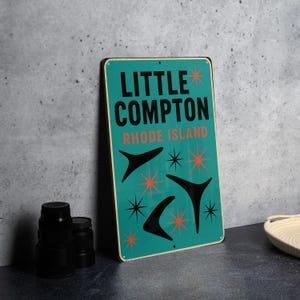 May include: A rectangular teal metal sign with the words "LITTLE COMPTON RHODE ISLAND" in black and orange. The sign features black boomerang and starburst designs. The sign has a vintage, retro aesthetic.