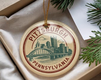 May include: A round ceramic Christmas ornament featuring a vintage-style illustration of the Pittsburgh, Pennsylvania skyline and the Fort Pitt Bridge. The ornament has a cream-colored background with a red and green design and the words "Pittsburgh Pennsylvania".