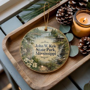 May include: A round ceramic ornament featuring a scenic landscape of John W. Kyle State Park, Mississippi. The ornament has a gold string for hanging. The image includes a waterfall, lake, trees, and the text "John W. Kyle State Park, Mississippi".