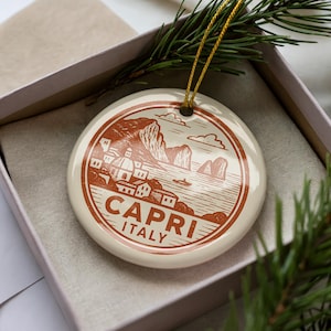May include: A round, off-white ceramic ornament featuring a brown illustration of Capri, Italy. The design includes buildings, cliffs, and the text "CAPRI ITALY". A gold string is attached for hanging. The ornament is presented in a gift box.