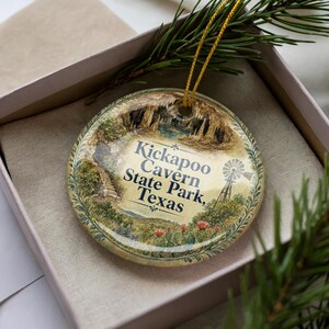 May include: A round Christmas ornament featuring a scenic view of Kickapoo Cavern State Park, Texas. The ornament has a gold string for hanging and is presented in a gift box. The image includes a windmill, cave, and text.