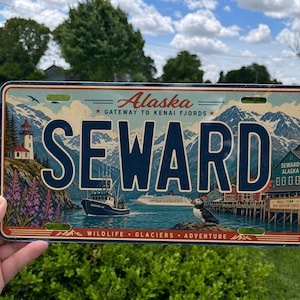 May include: A rectangular metal license plate featuring a scenic illustration of Seward, Alaska. The text reads Alaska, Gateway to Kenai Fjords, Seward, and Wildlife, Glaciers, Adventure. It depicts a lighthouse, mountains, a boat, and a puffin.