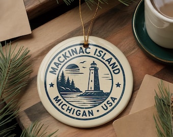 May include: Cream-colored ceramic ornament featuring a navy blue design of a lighthouse, trees, and the text "Mackinac Island Michigan USA". The ornament has a gold string for hanging and is a souvenir or decorative item.