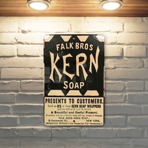 May include: A vintage metal sign advertising Kern Soap. The sign is black and white with red accents. It features the text "Falk Bros Kern Soap Presents to Customers." and instructions to send in 25 soap wrappers for a gift.