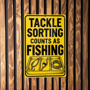 May include: A rectangular yellow metal sign with black text that reads TACKLE SORTING COUNTS AS FISHING. Below the text is a black line drawing of a three-compartment tray containing a fishing lure, a bobber, and a coiled fishing line.