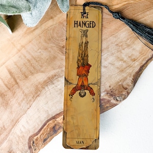 May include: A bookmark featuring a vintage-style illustration of The Hanged Man card from a tarot deck. The bookmark is printed on a parchment-like background with a black tassel.