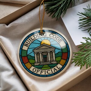May include: Round ceramic ornament with the words "BUILDING CODE OFFICIAL" around the edge. The center features a building with columns, a rising sun, and a stained-glass style design in blue, green, and orange.