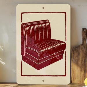 May include: A rectangular metal sign featuring a burgundy diner booth illustration. The booth is set against a cream background within a burgundy border. The sign has two mounting holes.