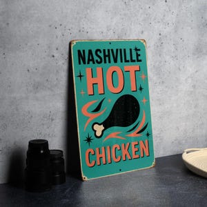 May include: A vintage-style metal sign with the words "Nashville Hot Chicken" in black and orange. The sign features a black chicken leg graphic with orange flames and starbursts on a teal background. The sign has a distressed look.