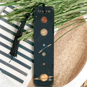 May include: A black bookmark with a gold tassel. The bookmark features a dark blue background with a stylized image of the solar system, including the sun, planets, and stars. The text "System" is printed in gold at the top of the bookmark.