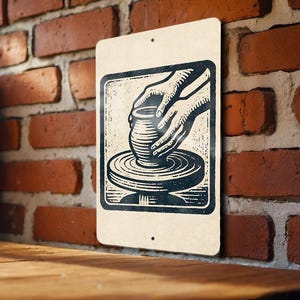 May include: A rectangular metal sign with a vintage illustration of hands shaping a pot on a pottery wheel. The artwork is in black and white, set against a cream-colored background. The sign is mounted on a brick wall.