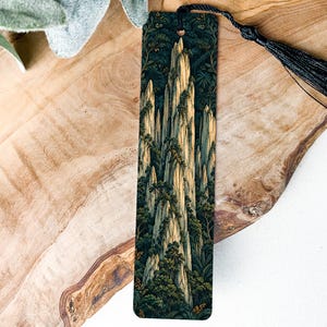 May include: A rectangular bookmark featuring a detailed illustration of tall, jagged mountain peaks surrounded by dense green foliage. The bookmark has a dark background and is finished with a black silk tassel attached to the top.