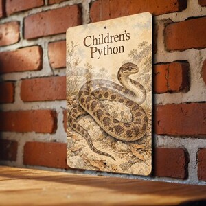 May include: A rectangular metal sign featuring a detailed illustration of a Children's Python snake coiled on a rock. The text Children's Python is printed in brown at the top. The background shows a natural landscape with dry vegetation.