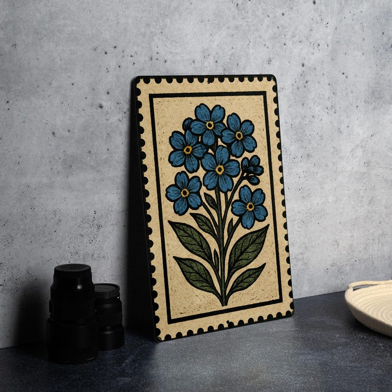 A framed artwork depicting a blue flower is displayed on a table, accompanied by a stack of black camera lenses and a white dish.