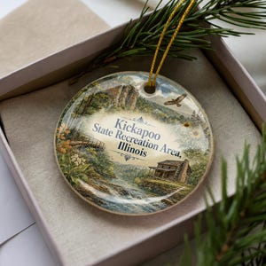 May include: A round ceramic ornament featuring a scenic landscape of Kickapoo State Recreation Area, Illinois. The ornament has a gold string for hanging and is presented in a gift box. The artwork depicts a river, bridge, and cabin.