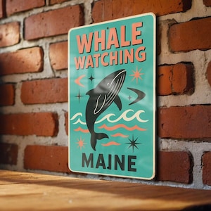 May include: A rectangular sign with the words "WHALE WATCHING MAINE" in coral lettering. The sign features a black whale illustration, waves, and starbursts on a teal background. The sign is displayed against a brick wall.
