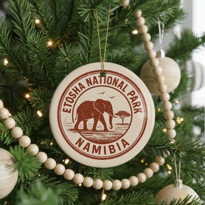 May include: A round, off-white Christmas ornament with a brown illustration of an elephant, tree, and text that reads "Etosha National Park Namibia". The ornament is hanging on a Christmas tree with other ornaments and wooden beads.