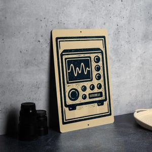 May include: A beige metal sign featuring a dark blue illustration of an oscilloscope. The oscilloscope has a screen displaying a waveform, with several knobs and buttons. The sign is rectangular with rounded corners and two holes for hanging.