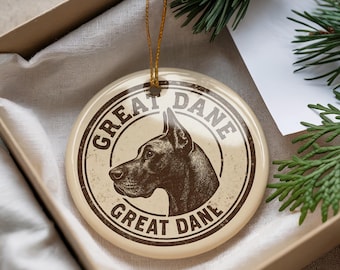 May include: A round, cream-colored Christmas ornament featuring a Great Dane dog illustration in a circular design. The words "GREAT DANE" are printed around the dog's profile in a vintage style. A gold string is attached for hanging.