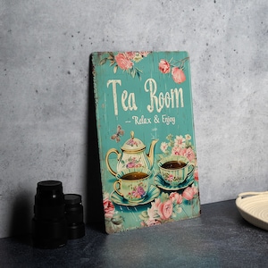 May include: A teal blue metal sign with a vintage design featuring a teapot and two teacups with floral patterns. The sign reads "Tea Room - Relax & Enjoy".