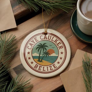 May include: A round, cream-colored ceramic ornament with a tropical beach scene featuring palm trees, a sunset, and the text "CAYE CAULKER BELIZE". The ornament has a gold string for hanging.
