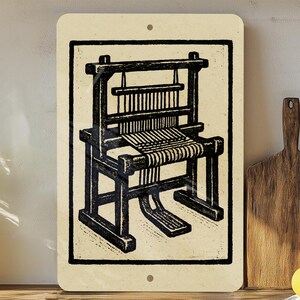 May include: A rectangular metal sign featuring a black and white illustration of a weaving loom. The loom is depicted in a vintage style, with intricate details of the frame, warp threads, and woven fabric. The sign has a cream-colored background.