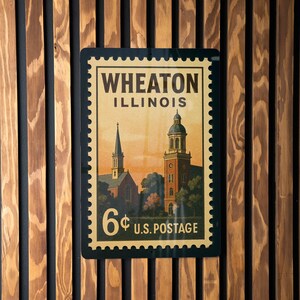 May include: A rectangular sign resembling a vintage postage stamp, featuring the text "WHEATON ILLINOIS" and a depiction of buildings. The sign reads "6¢ U.S. POSTAGE" and is set against a wooden slatted background.